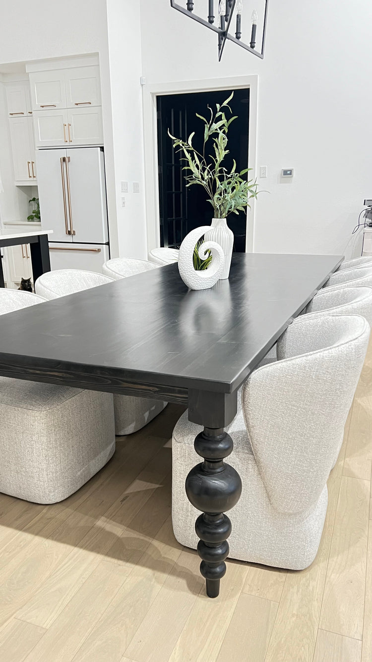 Large black rectangular table with curved pedestal legs, surrounded by eight large upholstered light gray chairs. A unique modern plant serves as the centerpiece, adding a touch of style to the space