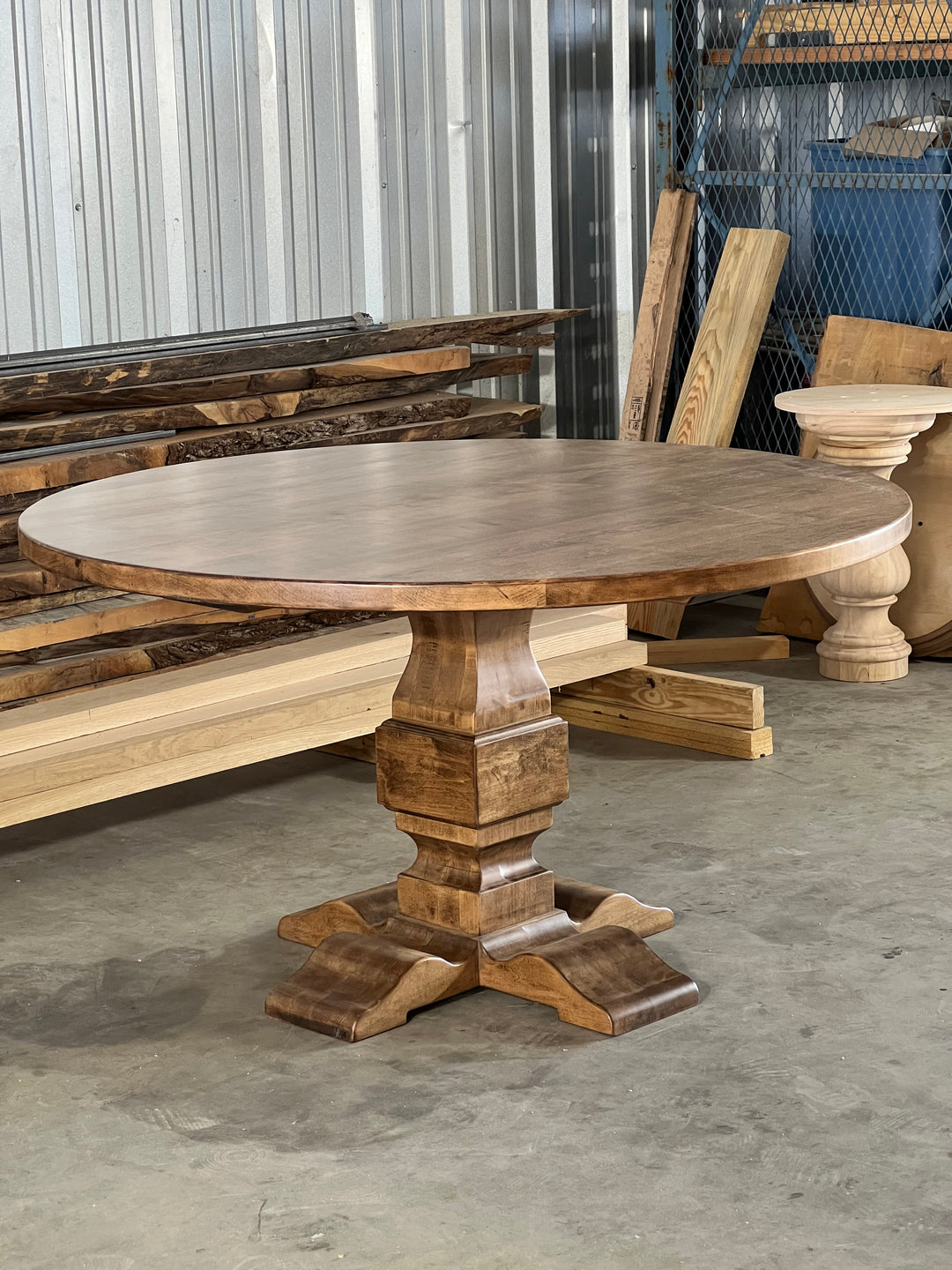 Warm brown round pedestal dining table in a woodworking shop, highlighting the smooth finish and detailed craftsmanship, with tools and wood materials in the background