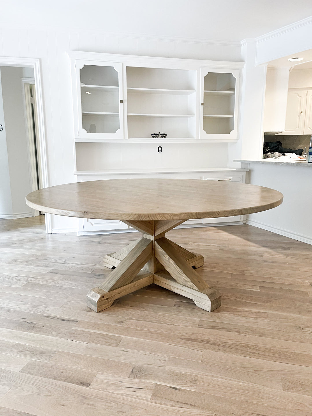 An expansive, round farmhouse dining table made of light brown pine, positioned in front of sleek white cabinets.