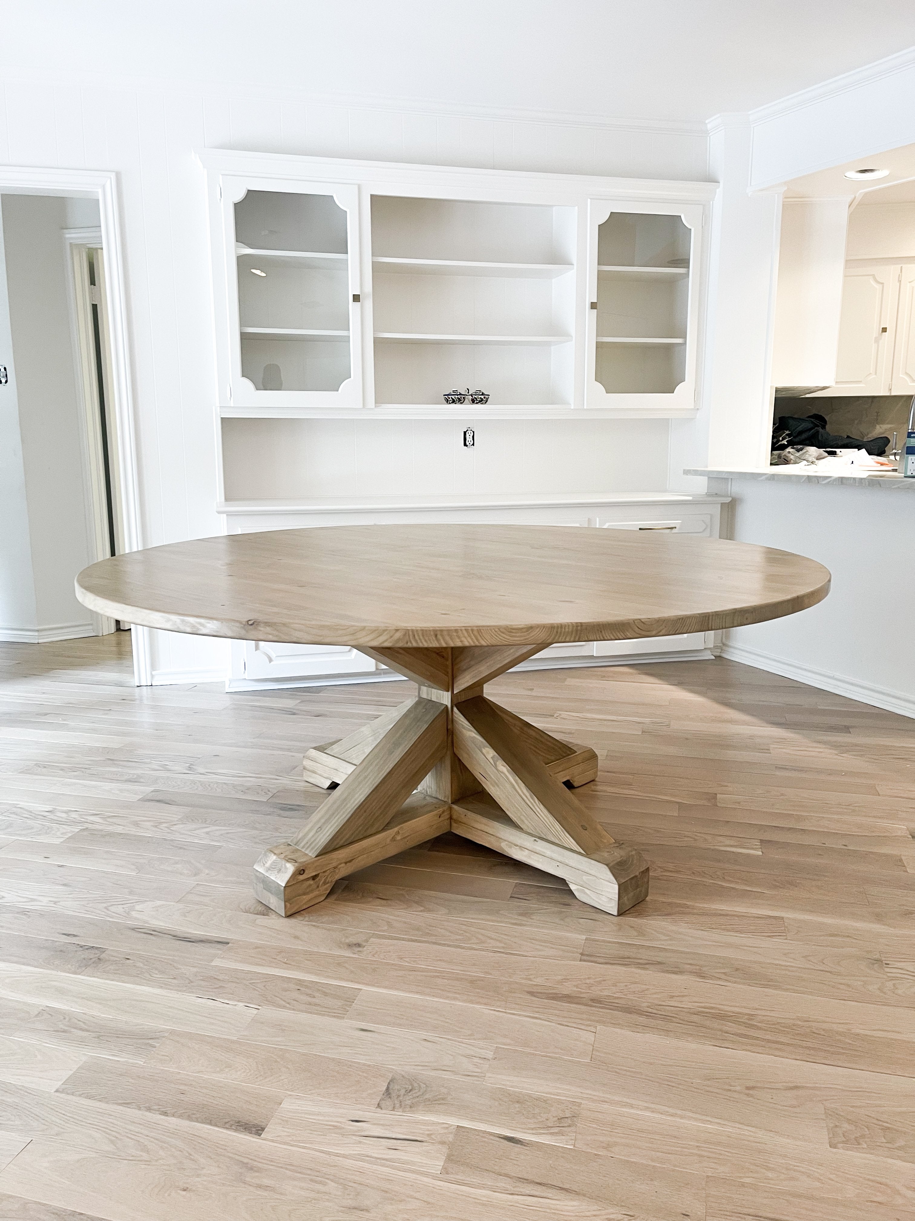 An expansive, round farmhouse dining table made of light brown pine, positioned in front of sleek white cabinets.