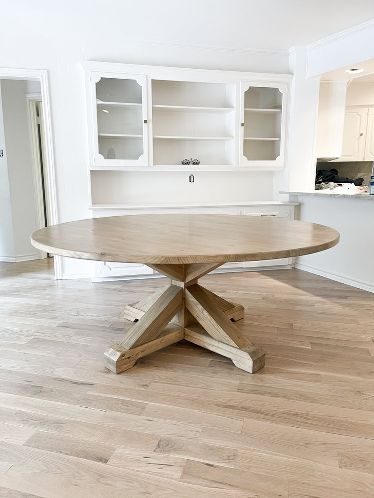 An expansive, round farmhouse dining table made of light brown pine, positioned in front of sleek white cabinets.