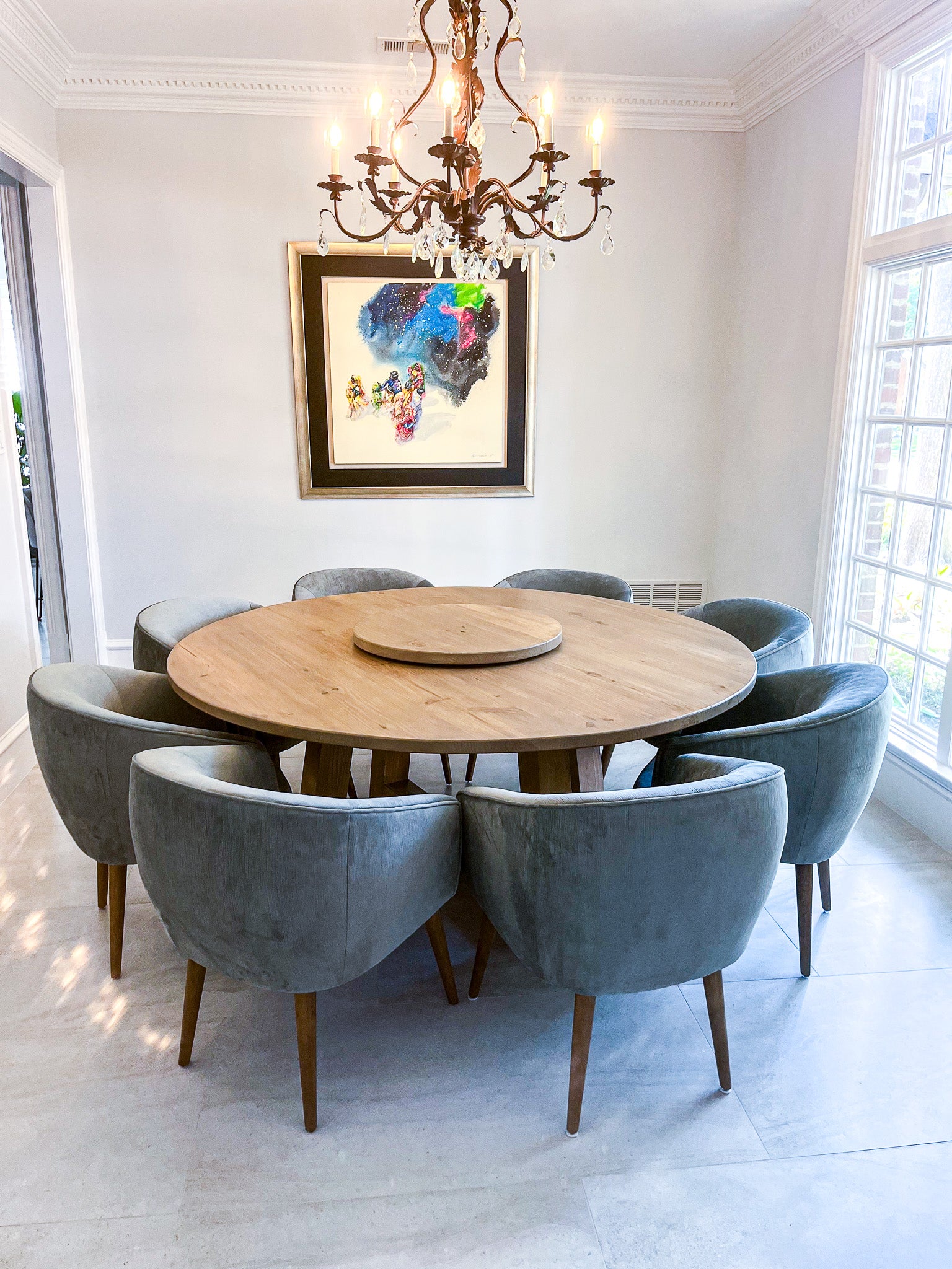 A round wooden dining table is situated in a bright Texas dining room, accompanied by eight wide upholstery chairs. The table features a trapezoid pedestal base, adding a striking modern farmhouse aesthetic.