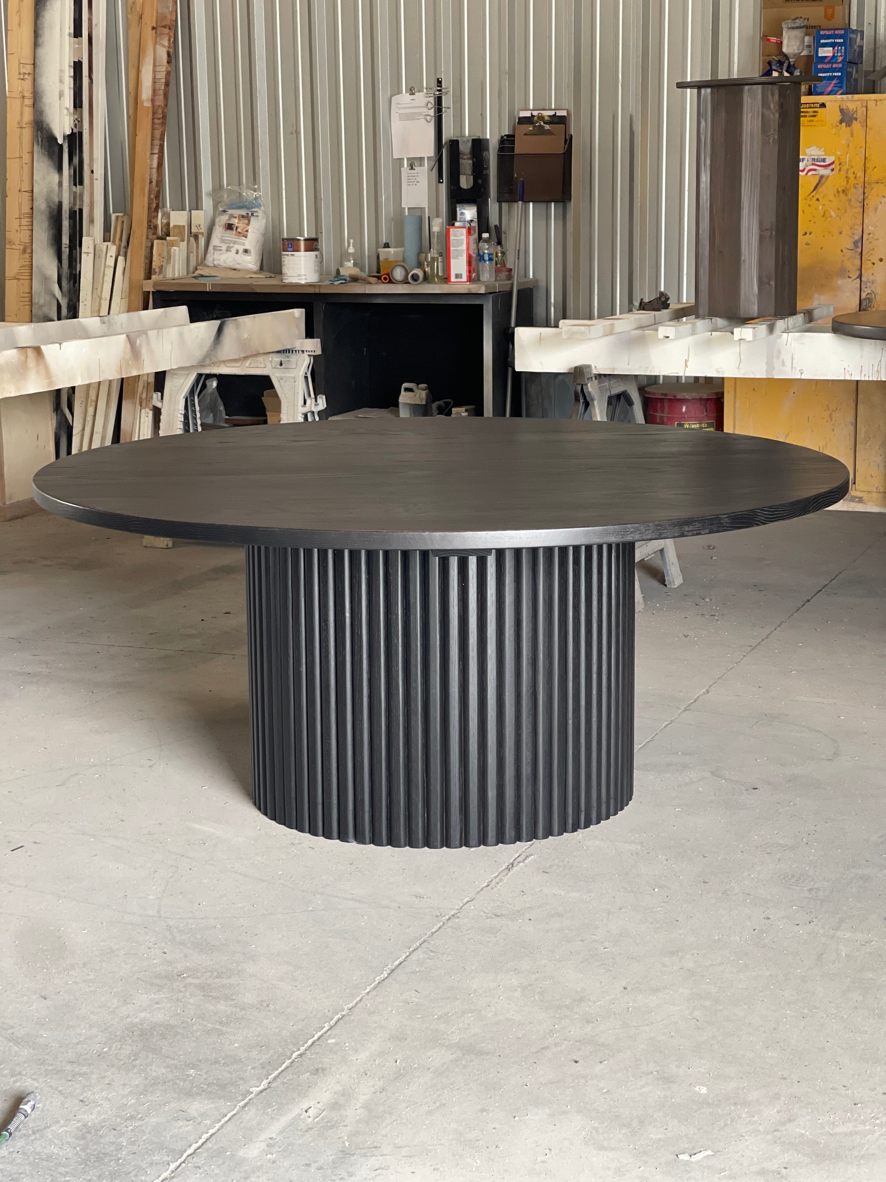 Large round black fluted dining table in a woodworking shop, with stains and paint cans in the background, reflecting the creative atmosphere of the space.