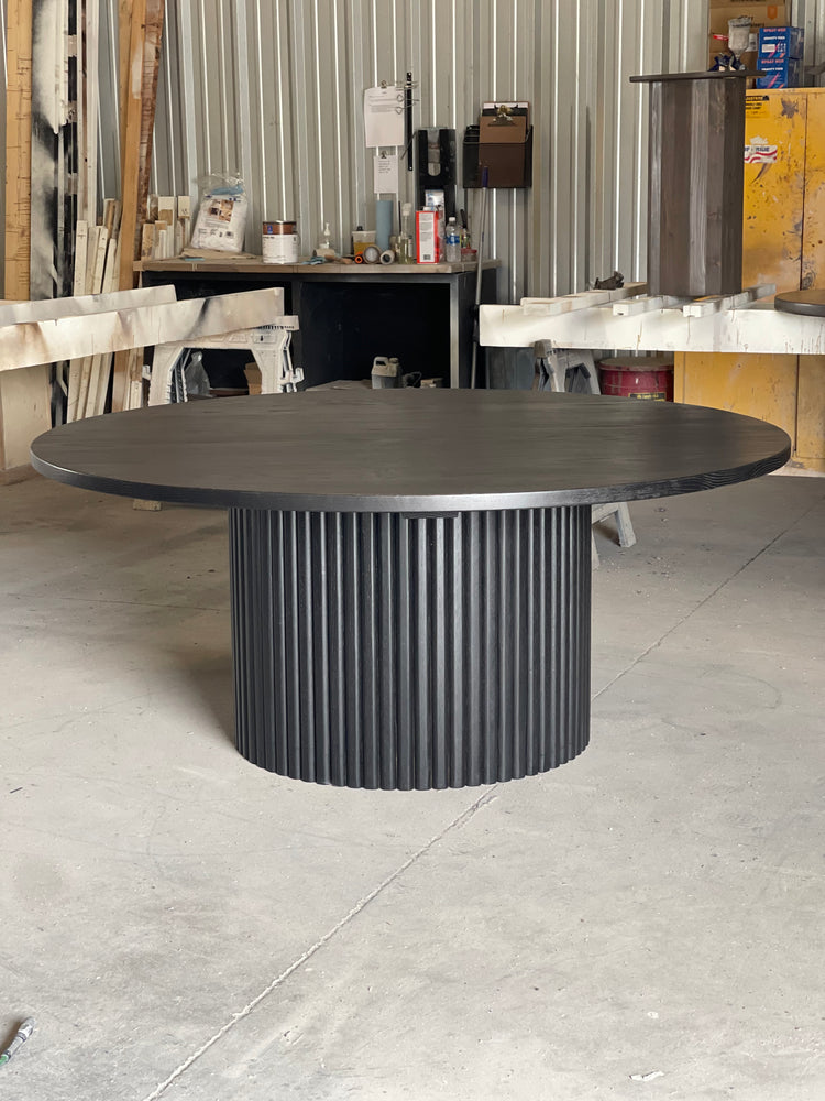 Large round black fluted dining table in a woodworking shop, with stains and paint cans in the background, reflecting the creative atmosphere of the space.