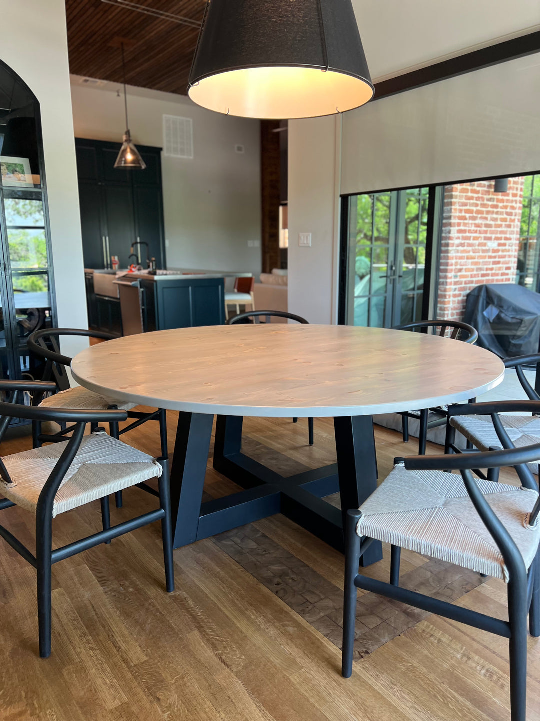 Round wooden dining table in a bright Texas dining room, set with 6 wide wishbone chairs. The table’s trapezoid black pedestal base creates an eye-catching modern farmhouse look.