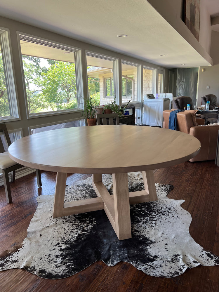 A large pale solid wood round dining table on a cow hide rug. The table features a trapezoid black pedestal base, adding a striking rustic farmhouse aesthetic