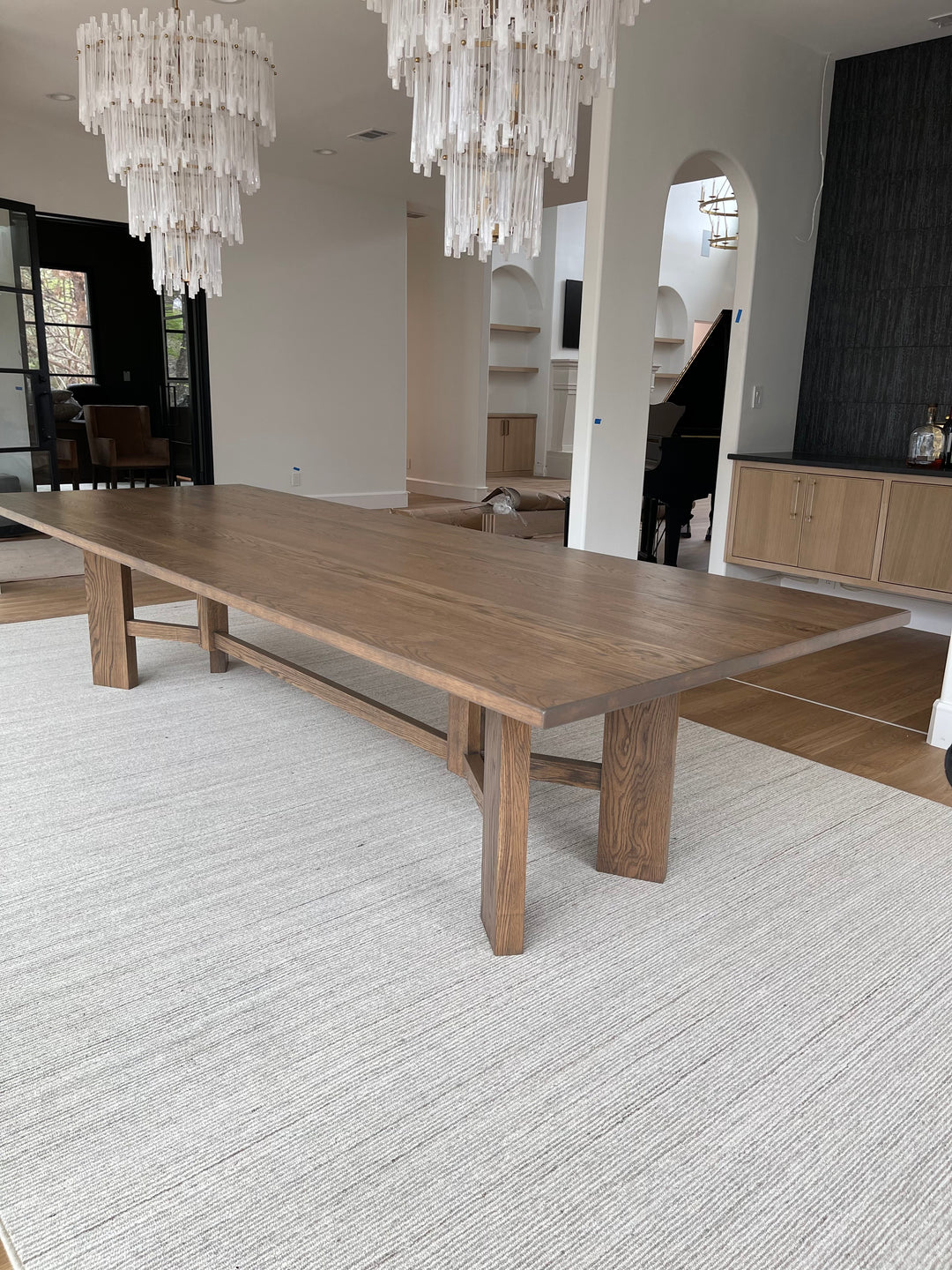 Large rectangular Briarsmoke white oak dining table in a grand and modern dining room, complemented by two elegant feather-like chandeliers and a gorgeous piano in the background, creating a luxurious and sophisticated atmosphere