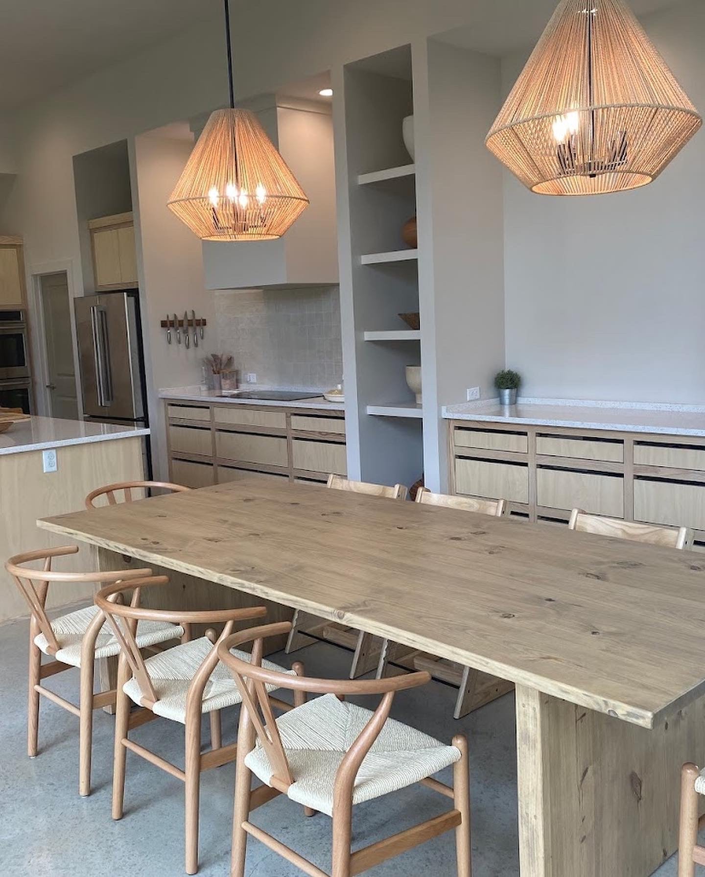 Light brown wooden rectangular dining table in a warm, cozy kitchen, surrounded by eight chairs, creating an inviting and family-friendly atmosphere. There are 2 wooden chandeliers hanging over. The ktichen has lots of wood accents