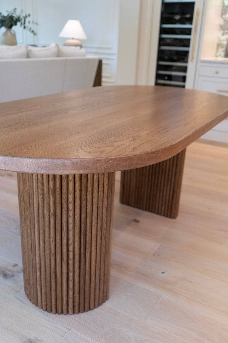 Wooden dining table with unique cylindrical legs in a room with a couch and lamp.