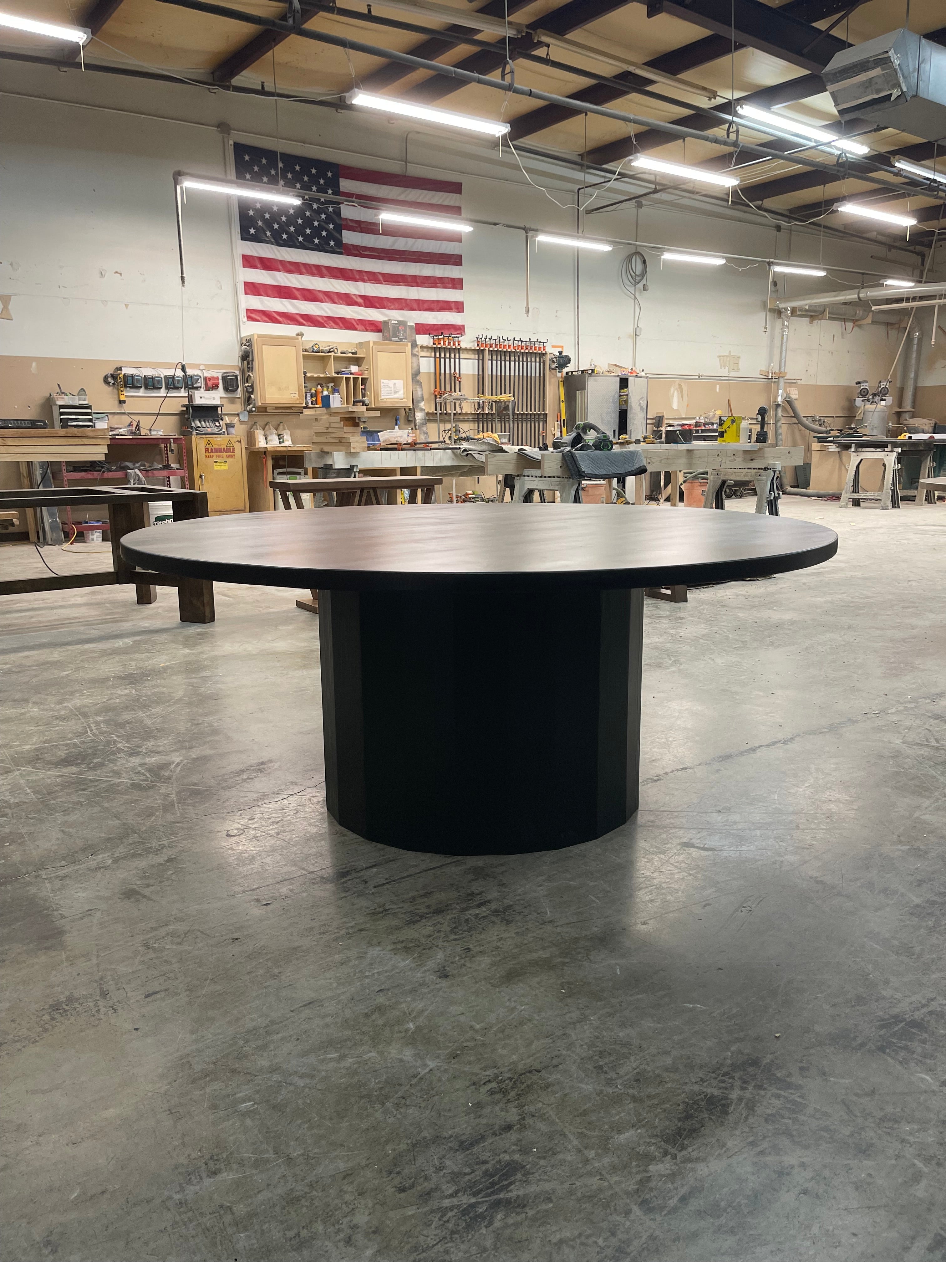 A large black round dining table situated in a woodworking warehouse in TX with an American flag in the background.
