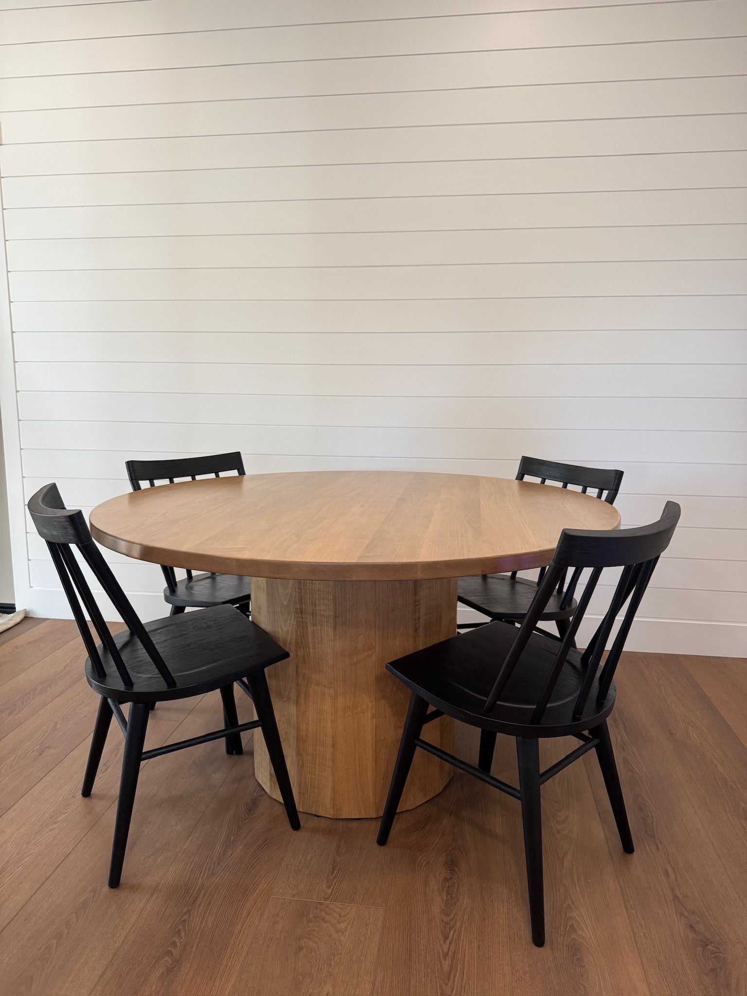 Warm cylindrical round dining table surrounded by four large black wooden chairs, set against a clean white background.