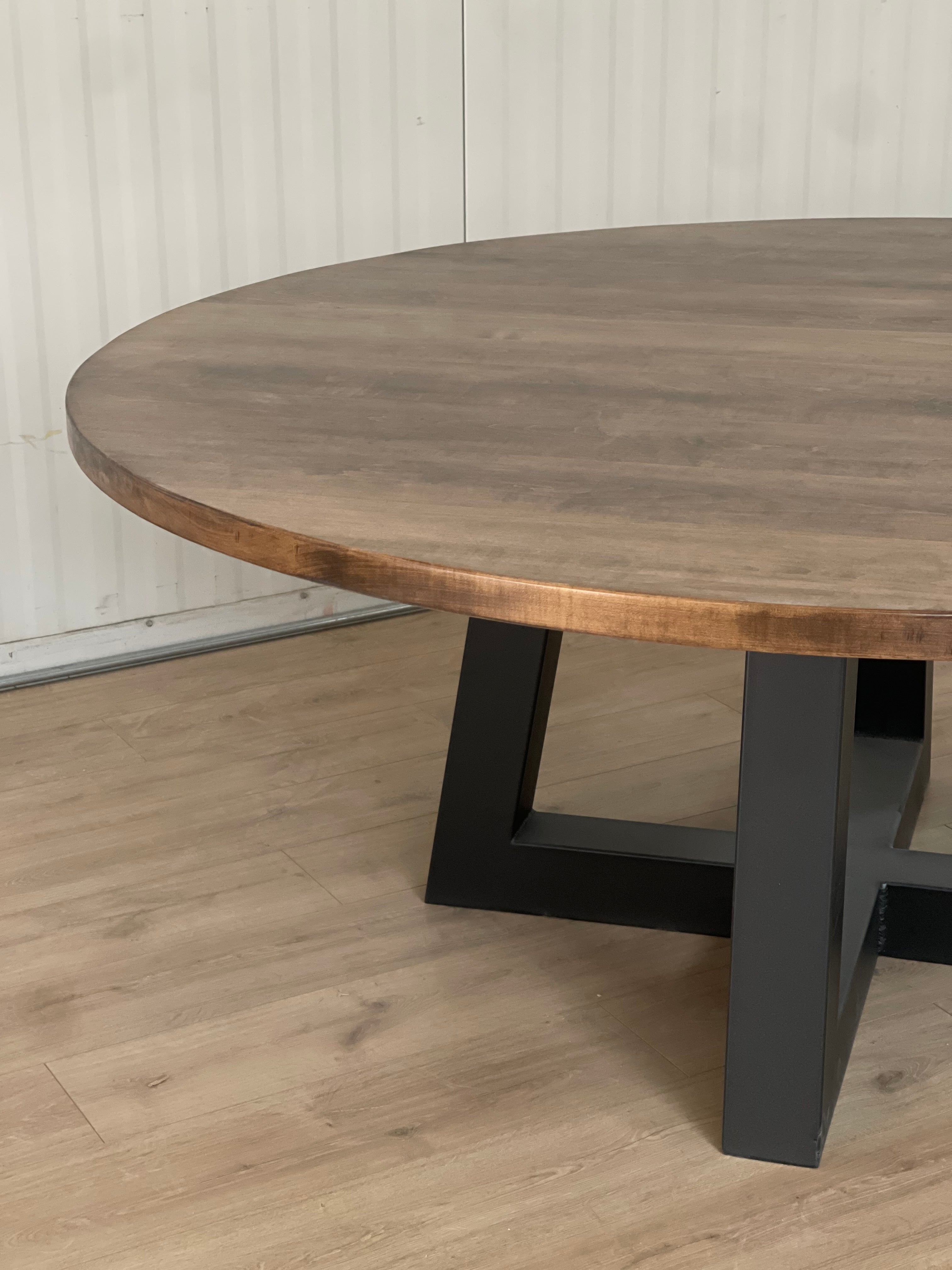Large maple round tabletop sitting on a black metal trapezoid base, creating a striking contrast between the warm wood and sleek, modern base