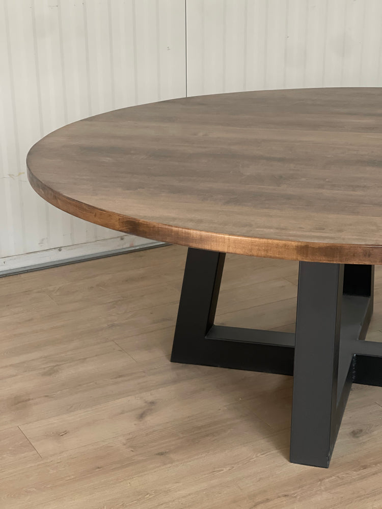 Large maple round tabletop sitting on a black metal trapezoid base, creating a striking contrast between the warm wood and sleek, modern base