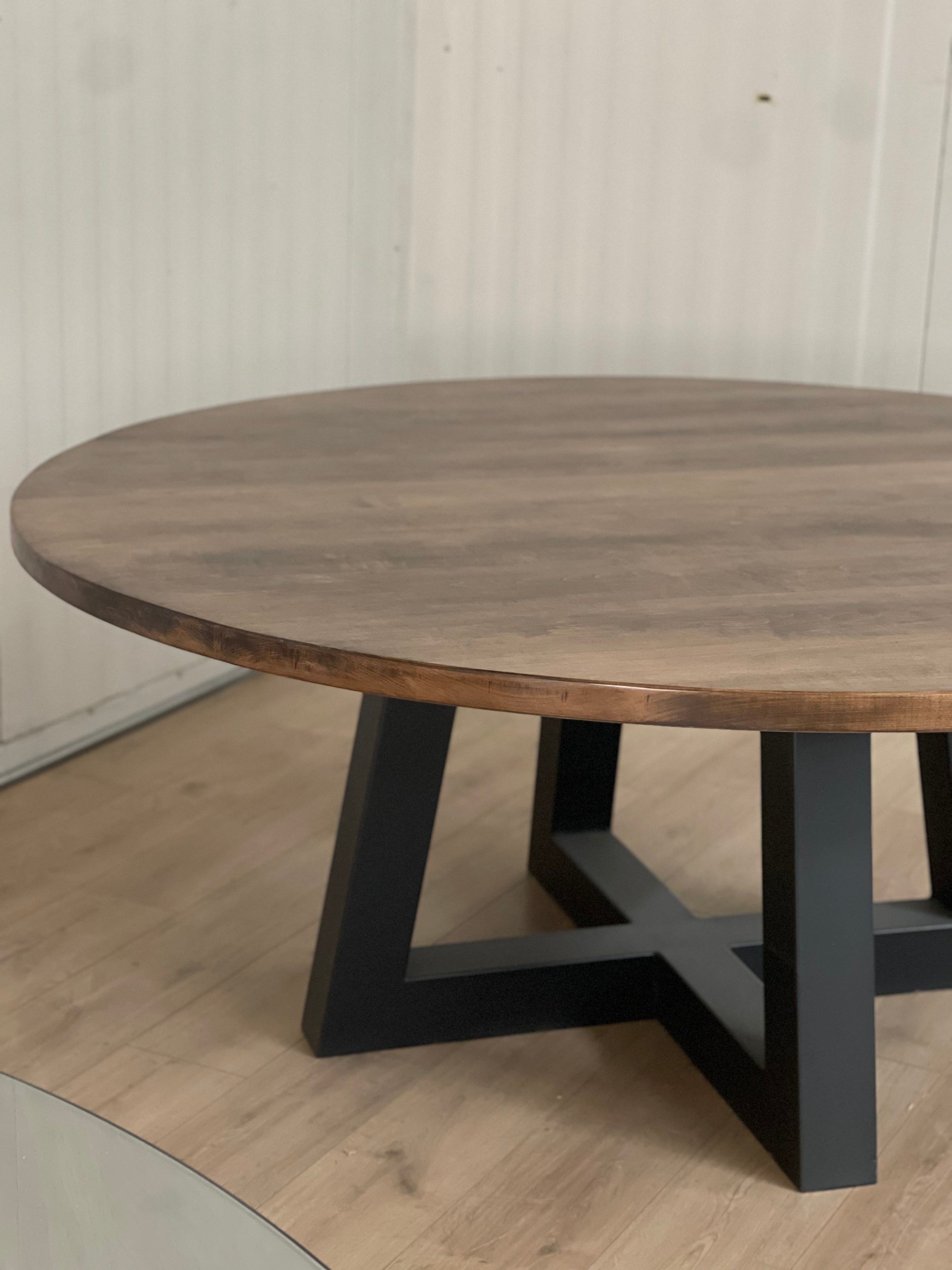 Large maple round tabletop sitting on a black metal trapezoid base, creating a striking contrast between the warm wood and sleek, modern base