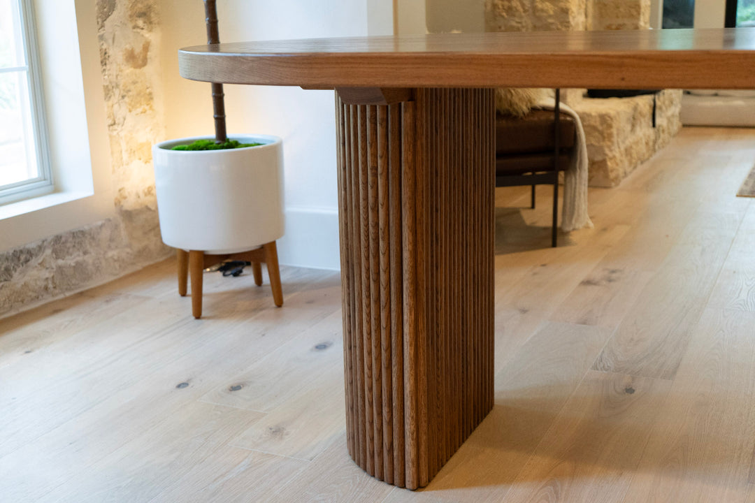 Wooden fluted white oak dining table with a textured column base in a room with stone walls and a plant.