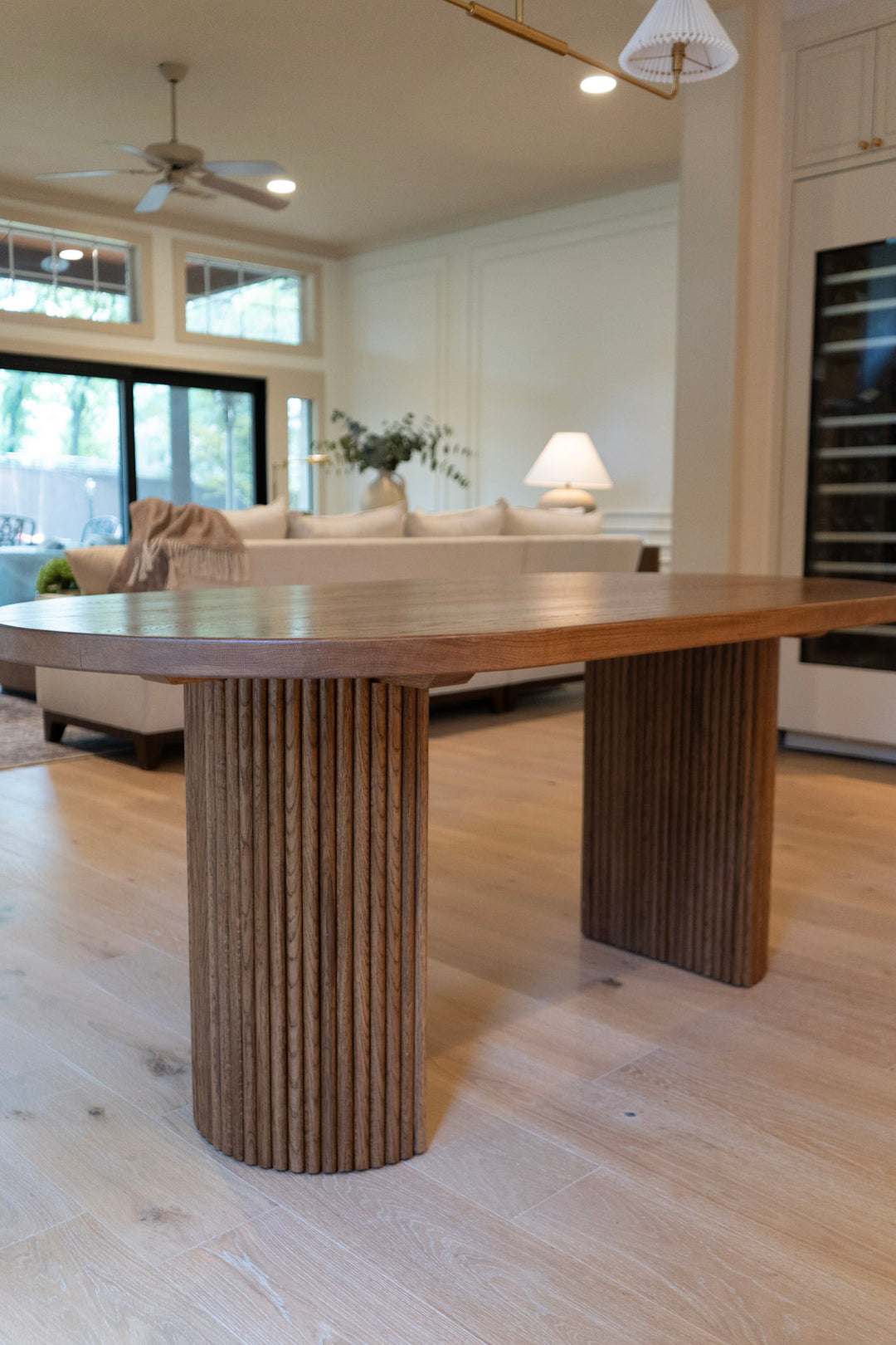 Wooden oval fluted dining table in a modern living room with a sofa and large windows.