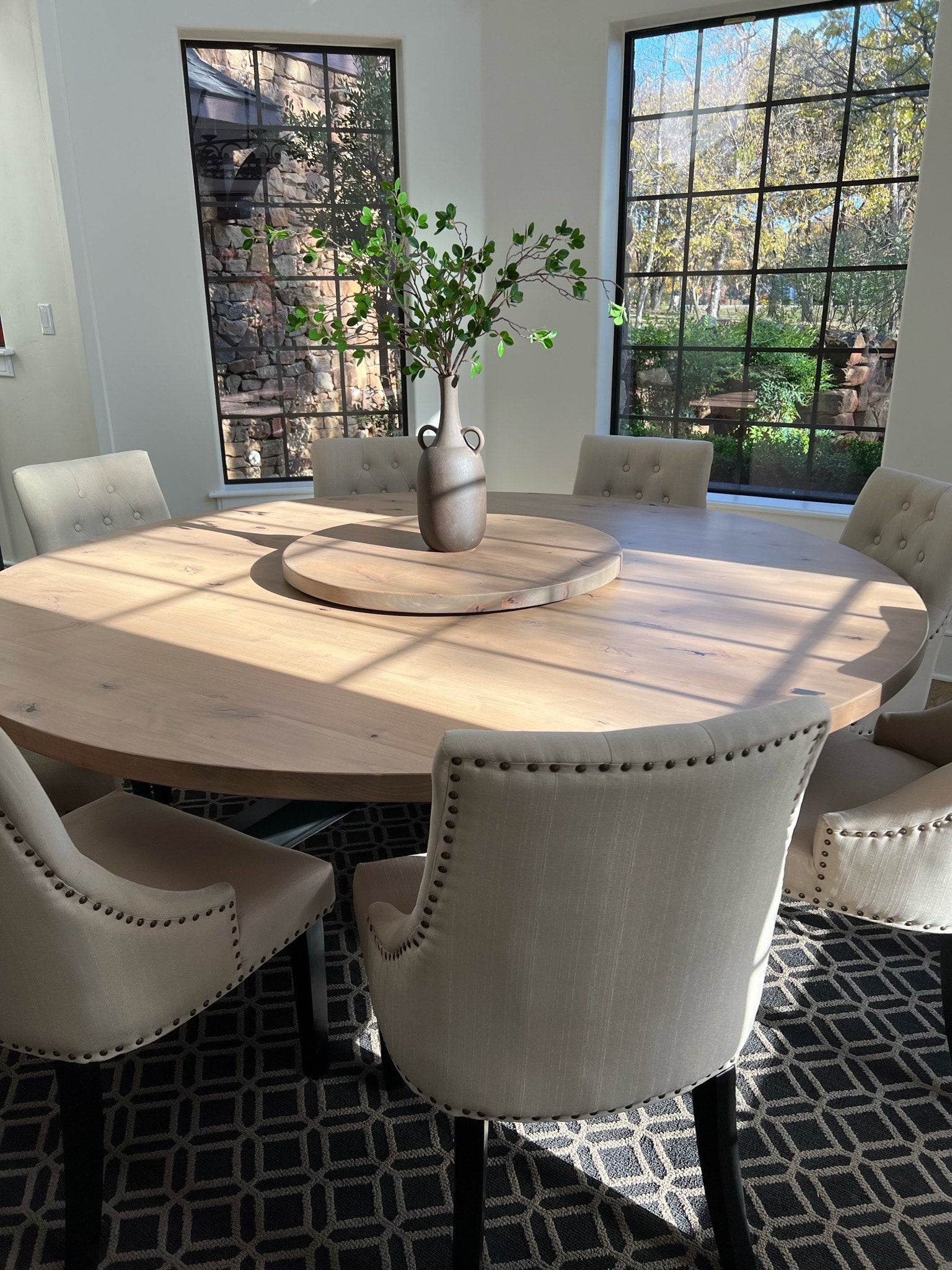 Large modern round wooden dining table surrounded by eight oversized beige upholstered chairs, with a lazy Susan and a plant as the centerpiece. The table is bathed in natural light from surrounding windows.