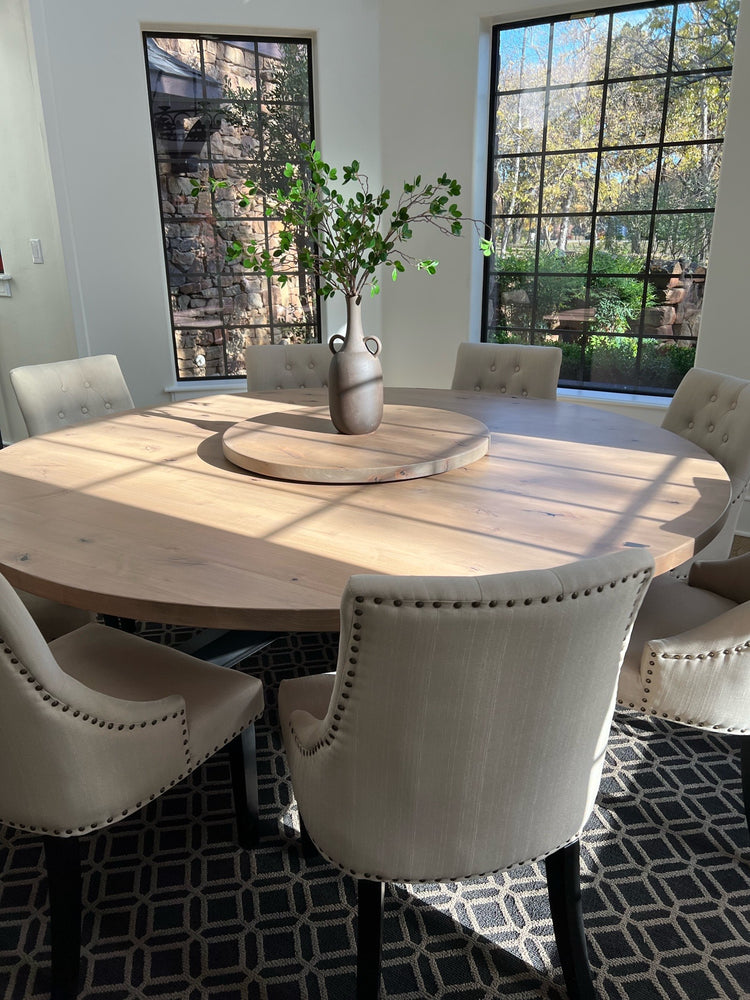 Large modern round wooden dining table surrounded by eight oversized beige upholstered chairs, with a lazy Susan and a plant as the centerpiece. The table is bathed in natural light from surrounding windows.