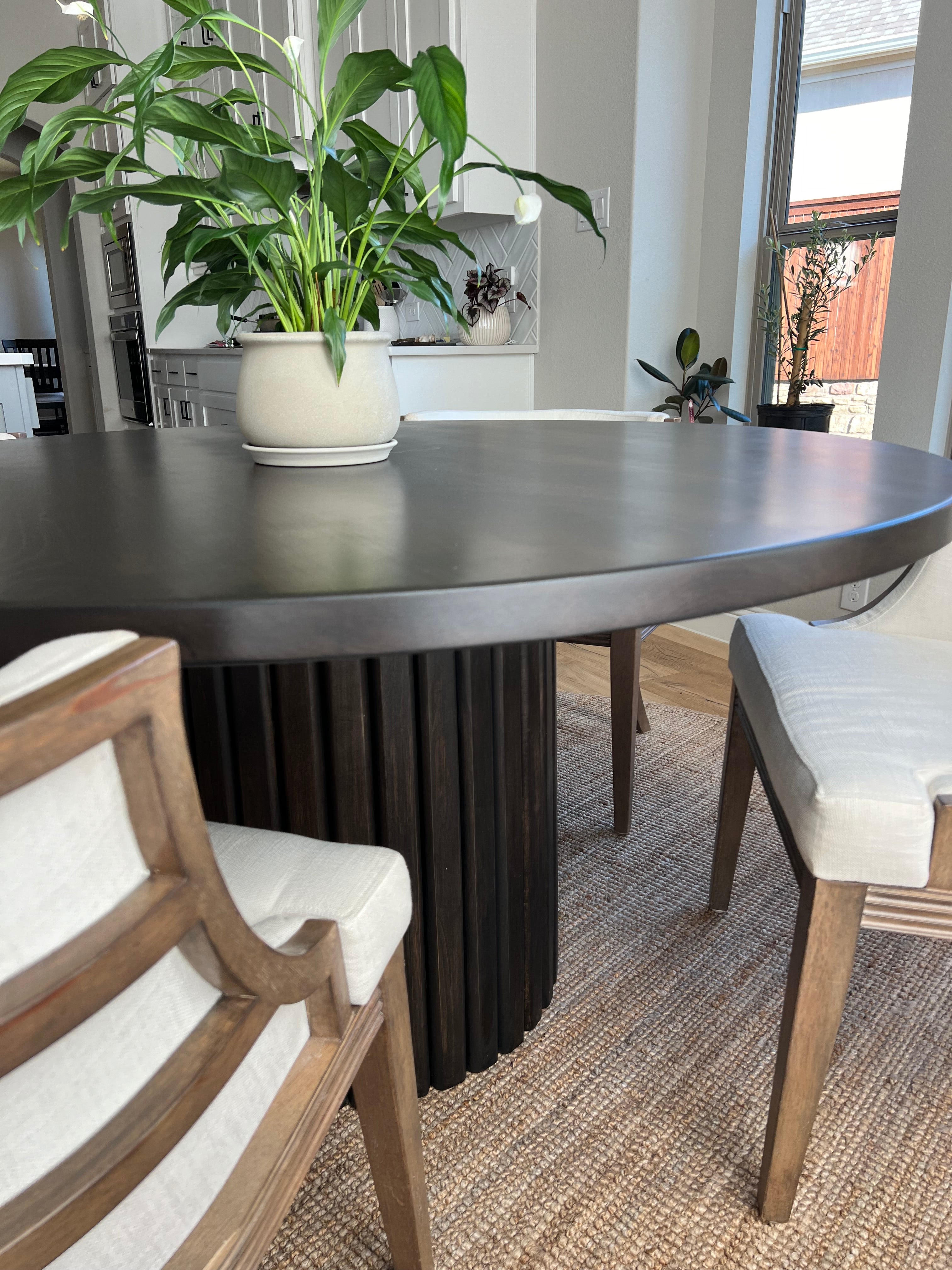 Up-close view of an onyx-colored round fluted dining table, surrounded by two large chairs and featuring a green plant as the centerpiece. The table is placed on a woven rug, adding texture to the space