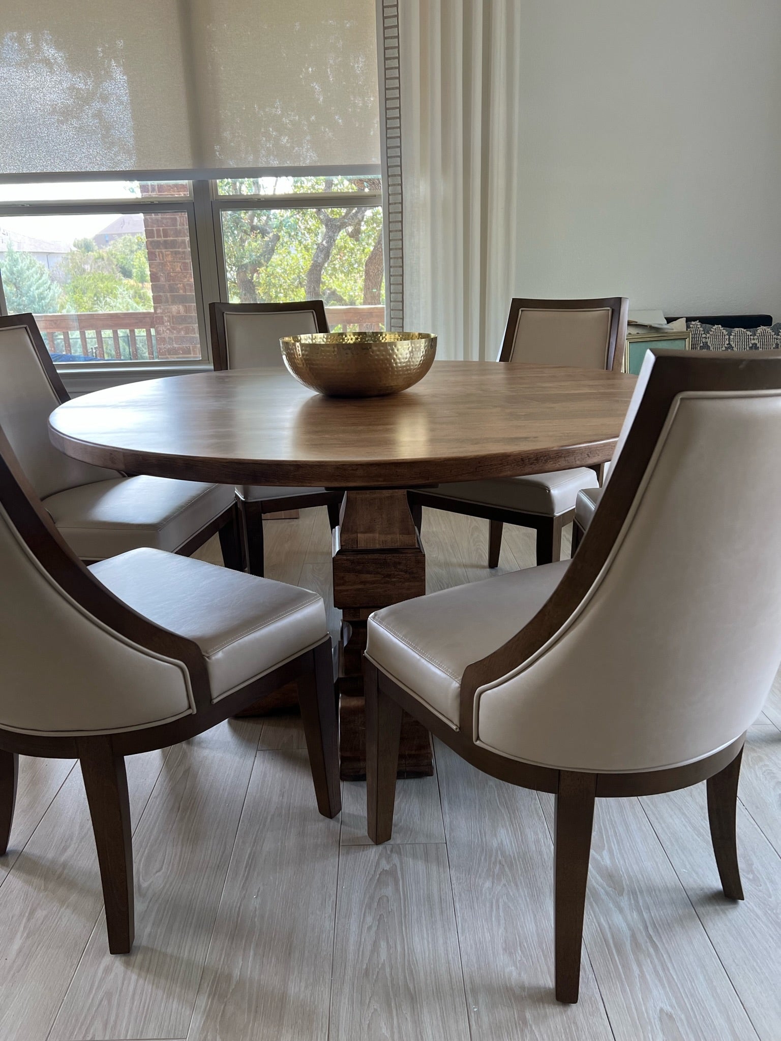 Warm brown round pedestal dining table in a modern, classy dining room, surrounded by six large cream leather chairs and a gold fruit bowl in the middle, creating an elegant and inviting atmosphere