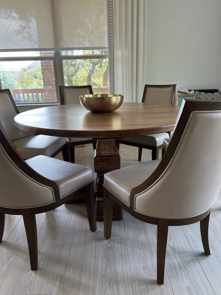 Warm brown round pedestal dining table in a modern, classy dining room, surrounded by six large cream leather chairs and a gold fruit bowl in the middle, creating an elegant and inviting atmosphere
