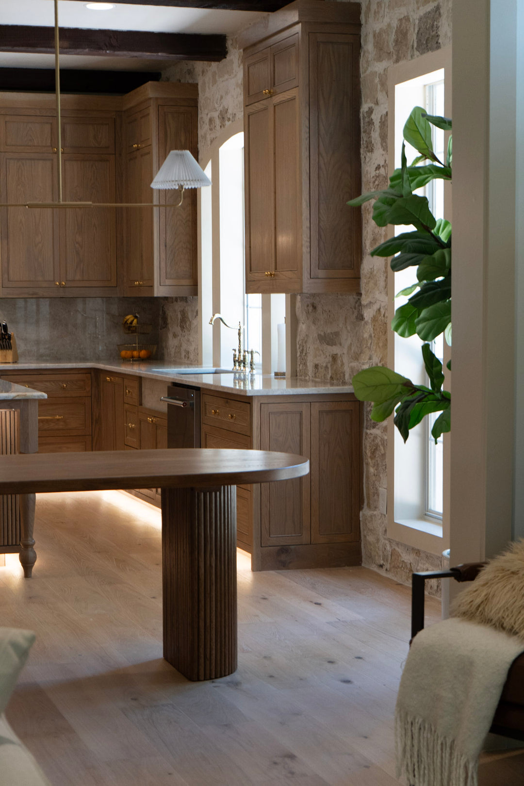 Modern fluted white oak dining table with oval top and half-cylinder base in front of Modern kitchen with wooden cabinets, a stone wall, and a plant.