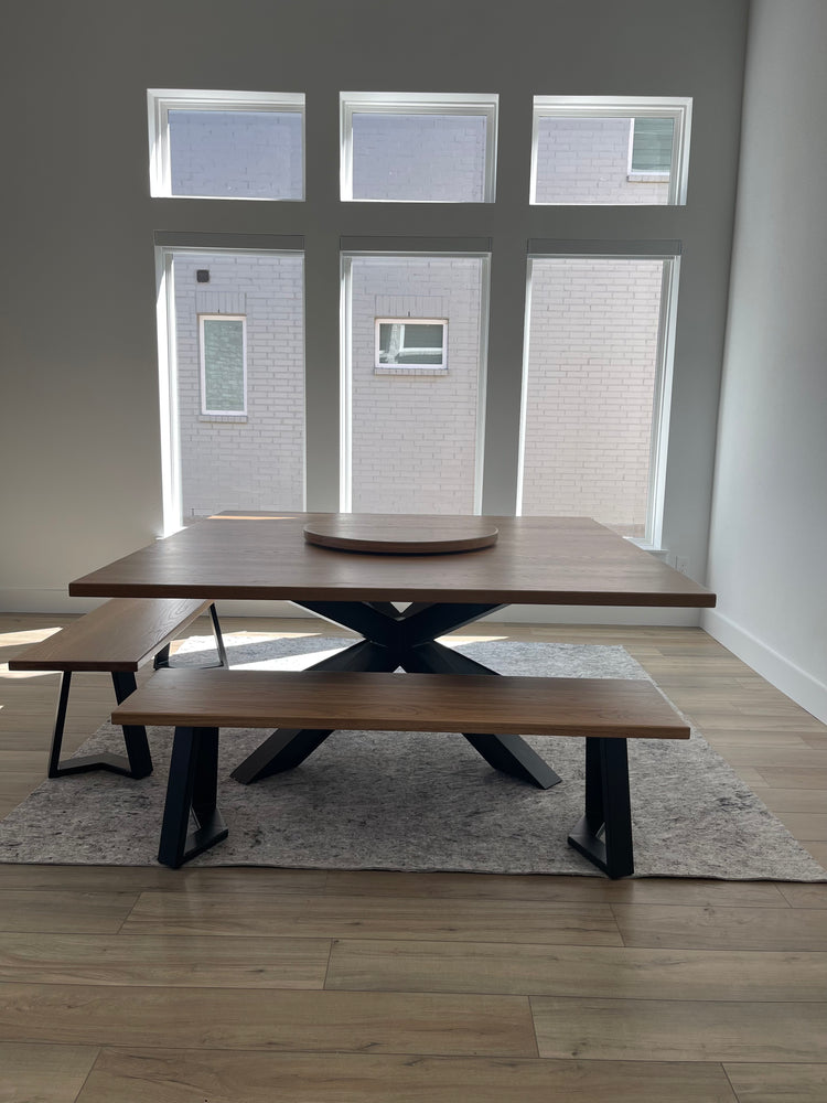 Large square metal dining table paired with two matching metal benches, featuring a lazy Susan on top. The table is positioned in front of a window, enhancing the modern and industrial aesthetic