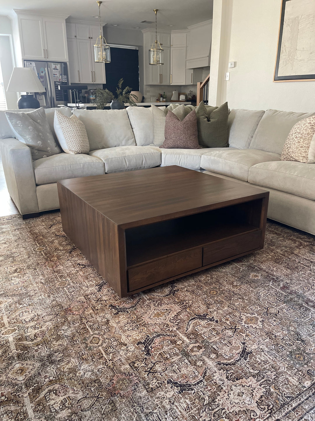 Dark brown rectangular wood coffee table with four front-facing drawers, placed on a red living room rug and surrounded by a large white sectional couch with decorative pillows