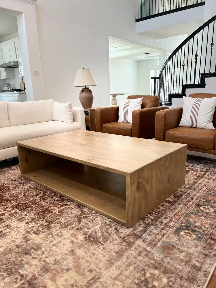 Sculptural donut-shaped coffee table centered on a muted red patterned rug, framed by a white couch and two rich brown leather armchairs with accent pillows—offering a balanced blend of modern form and classic warmth