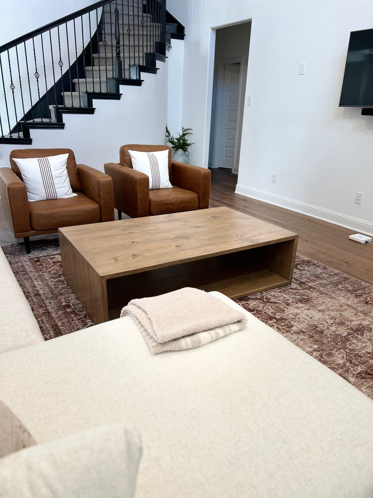 Light brown coffee table viewed from the kitchen, placed in front of two large leather armchairs on a red patterned rug, with graceful curved stairs rising in the background—creating a warm, layered living space