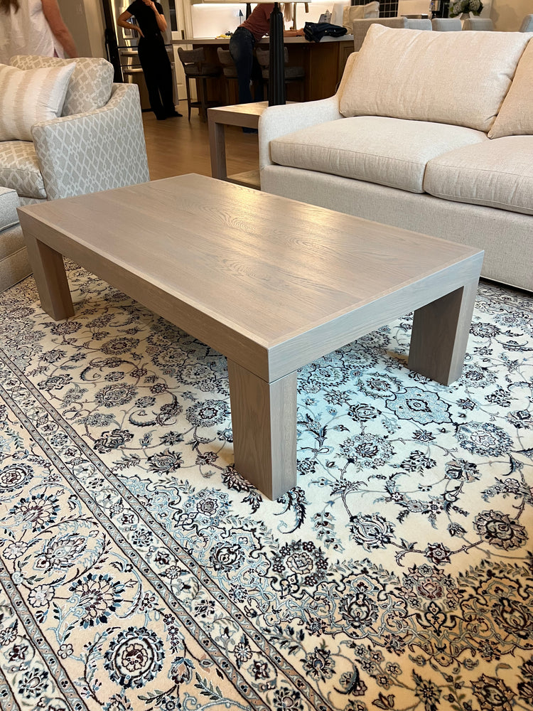 Custom white oak modern coffee table with thick block legs and a light neutral finish, placed on a patterned rug in a contemporary living room with built-in shelves and neutral-toned furniture.