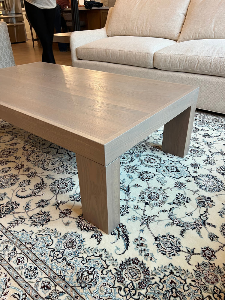 Custom white oak modern coffee table with thick block legs and a light neutral finish, placed on a patterned rug in a contemporary living room with built-in shelves and neutral-toned furniture.