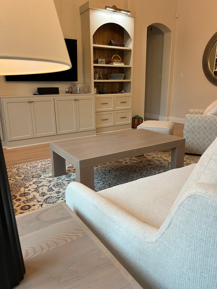 Custom white oak modern coffee table with thick block legs and a light neutral finish, placed on a patterned rug in a contemporary living room with built-in shelves and neutral-toned furniture.