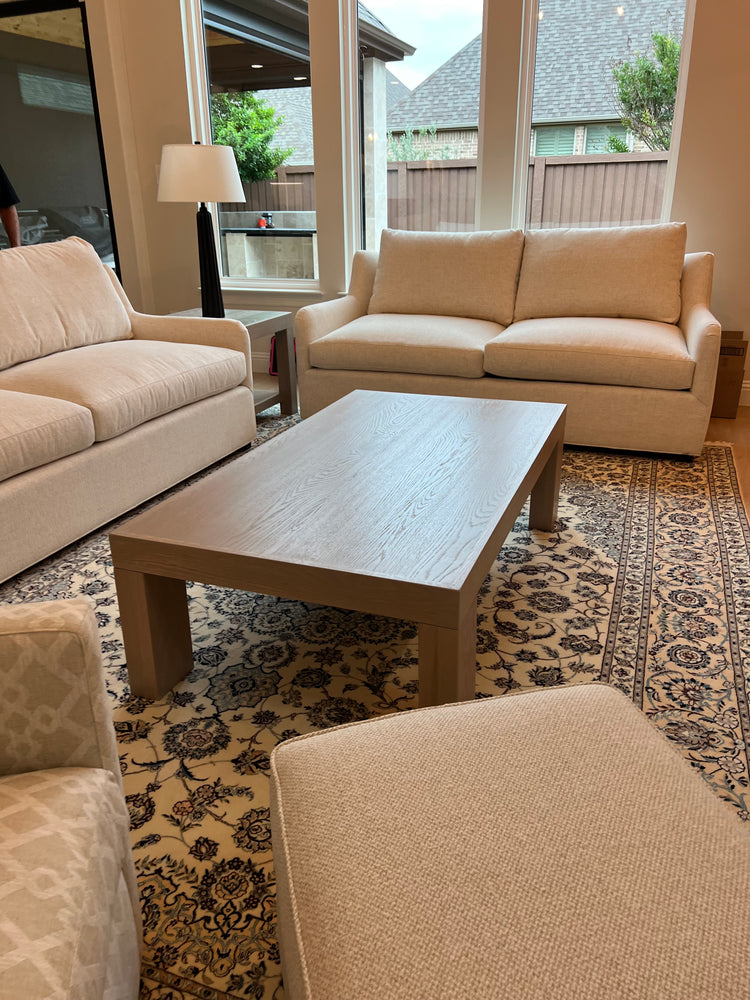 Custom white oak modern coffee table with thick block legs and a light neutral finish, placed on a patterned rug in a contemporary living room with built-in shelves and neutral-toned furniture.