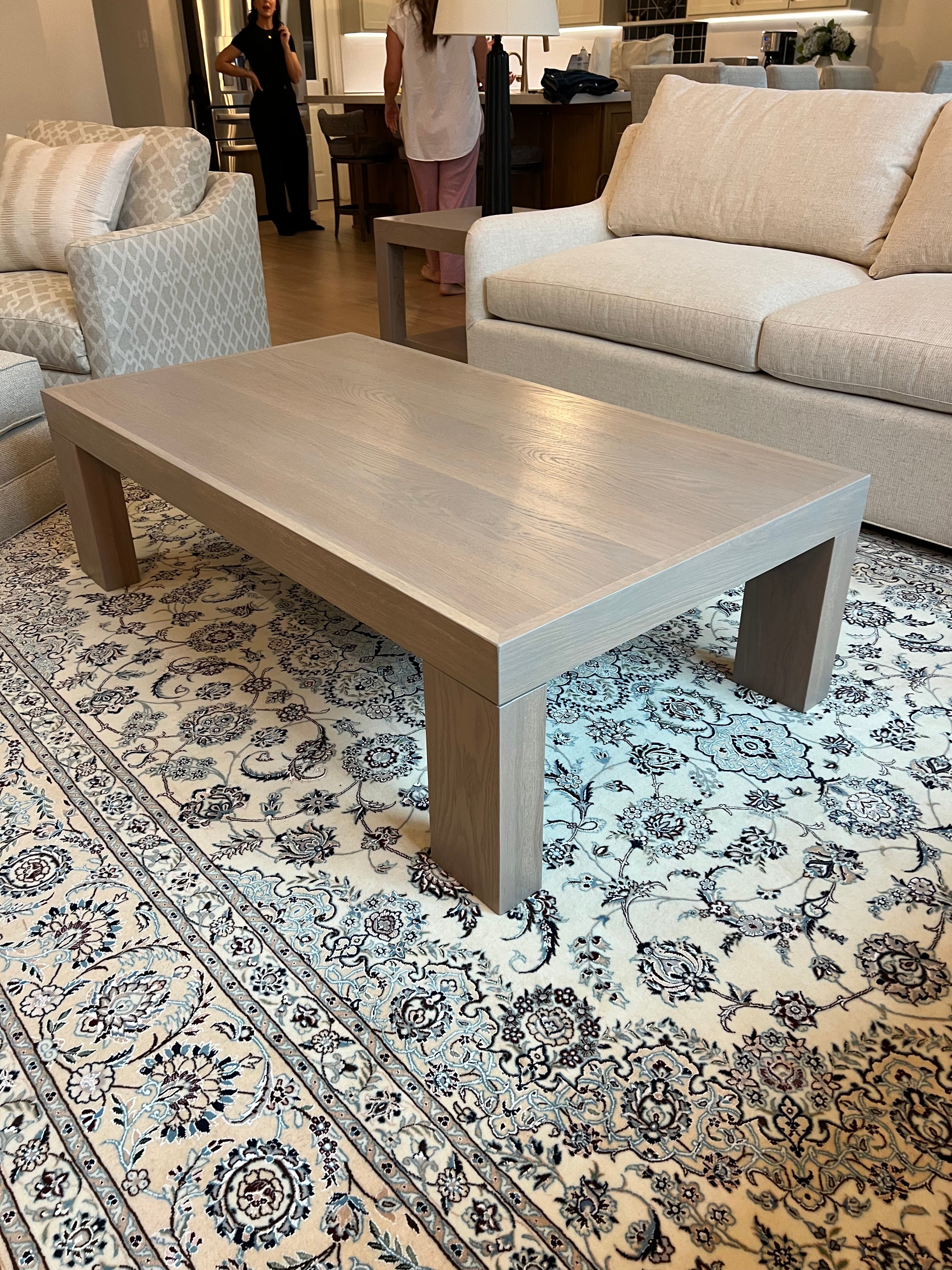 Custom white oak modern coffee table with thick block legs and a light neutral finish, placed on a patterned rug in a contemporary living room with built-in shelves and neutral-toned furniture.