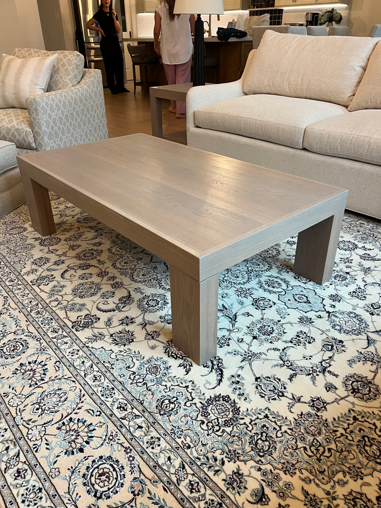 Custom white oak modern coffee table with thick block legs and a light neutral finish, placed on a patterned rug in a contemporary living room with built-in shelves and neutral-toned furniture.