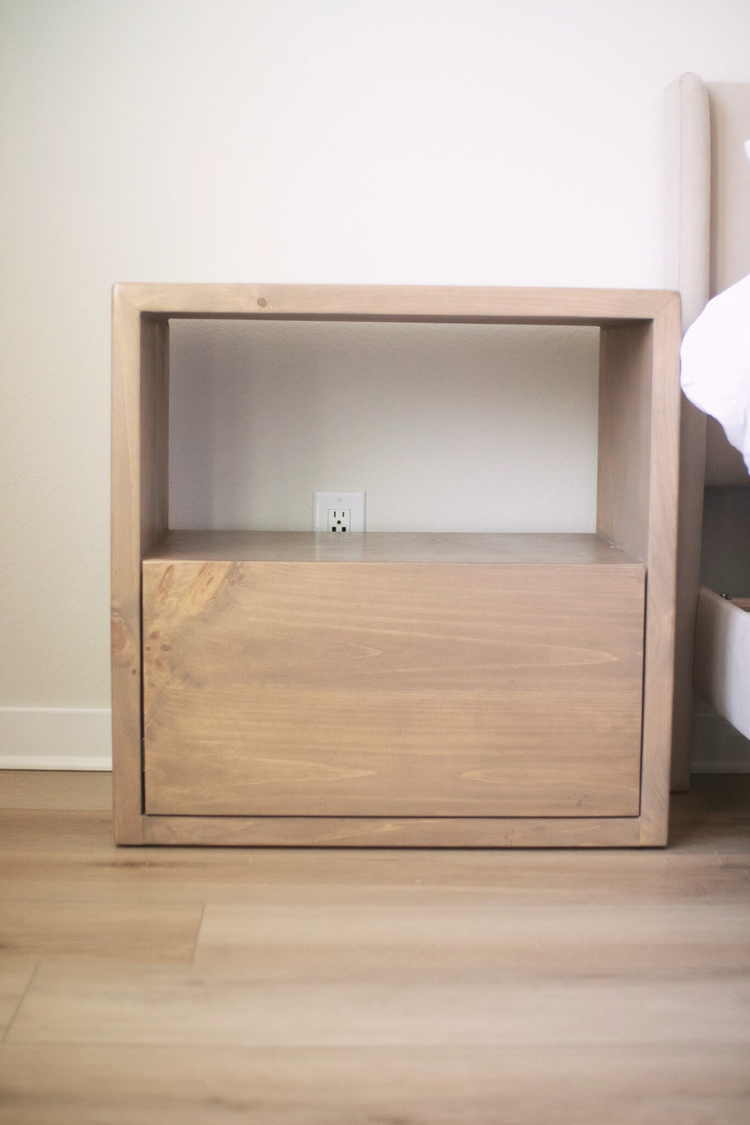 Front view of a ight brown nightstand positioned beside a white bed, creating a warm, natural contrast in a serene and minimalist bedroom setting