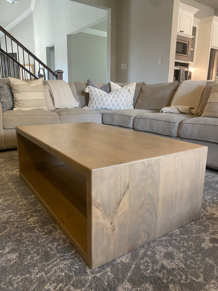Light brown coffee table nestled within a large gray sectional sofa adorned with pillows, all set atop a soft gray rug—creating a cozy, cohesive lounge area with warm natural tones